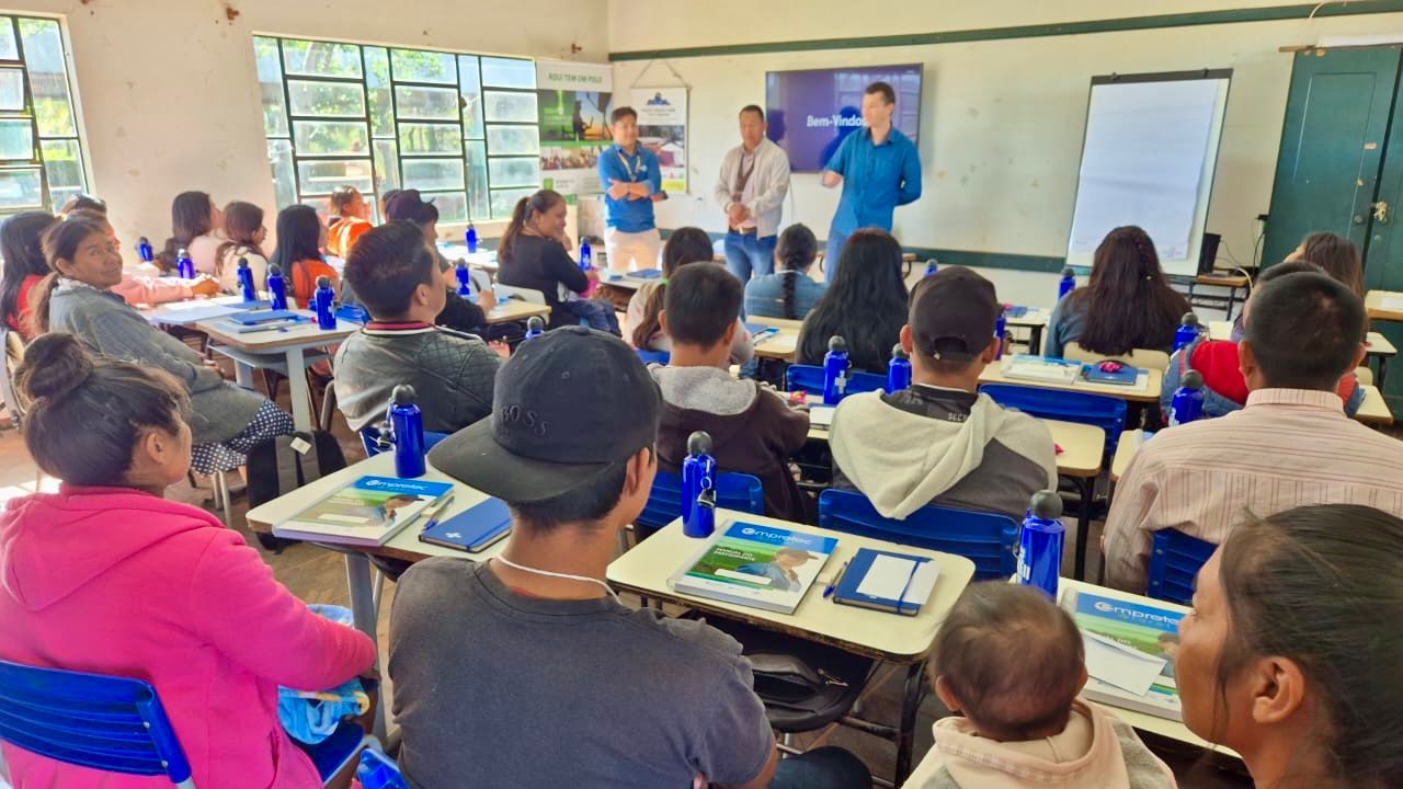 Em uma sala de aula iluminada, grupo de pessoas indígenas participa de uma capacitação do Sebrae, com materiais sobre as carteiras. Três facilitadores estão em pé à frente, falando ao grupo diante de uma tela com a palavra ‘Bem-vindos’.