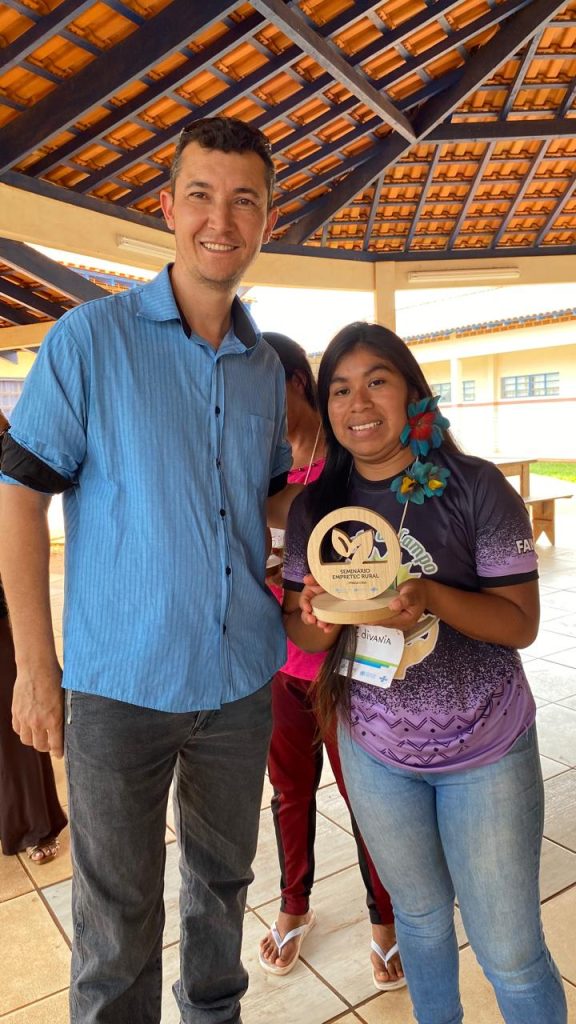 Um homem de camisa azul e uma mulher indígena sorridente com uma camiseta roxa e jeans posam lado a lado, segurando um prêmio de madeira redondo que diz 'Seminário Empretec Rural