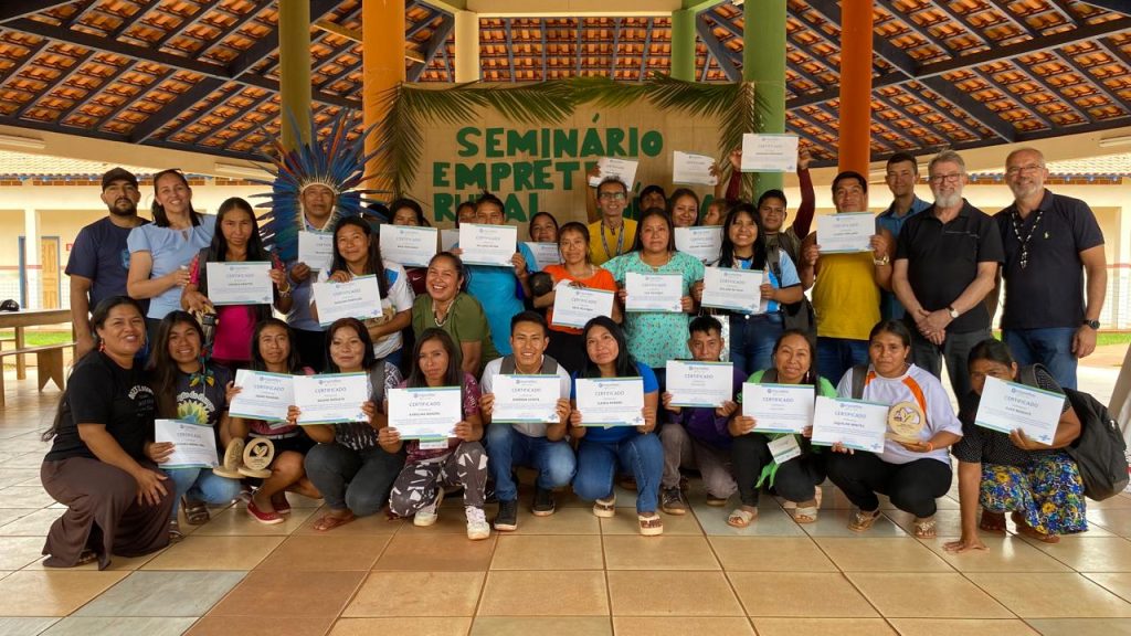 Grupo de participantes indígenas posa para foto com certificados nas mãos após o Seminário Empretec Rural Indígena, em Paranhos. Ao fundo, faixa com o nome do evento e uma estrutura colorida com colunas e telhado de madeira. Todos sorriem, celebrando a conclusão da capacitação