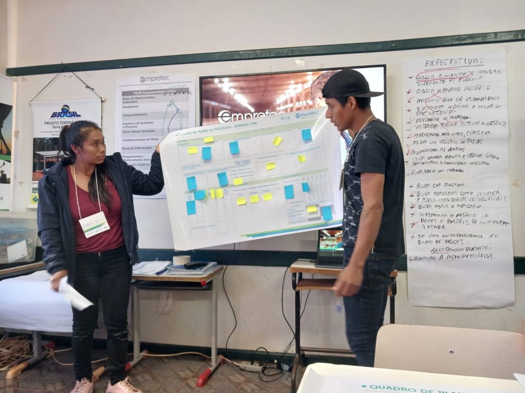 Dois participantes indígenas apresentam o quadro de plano de negócios durante atividade do Empretec Rural Indígena. Eles seguram uma cartolina com anotações e post-its coloridos, enquanto um painel ao fundo mostra o logotipo do programa e um cartaz exibe as expectativas do grupo