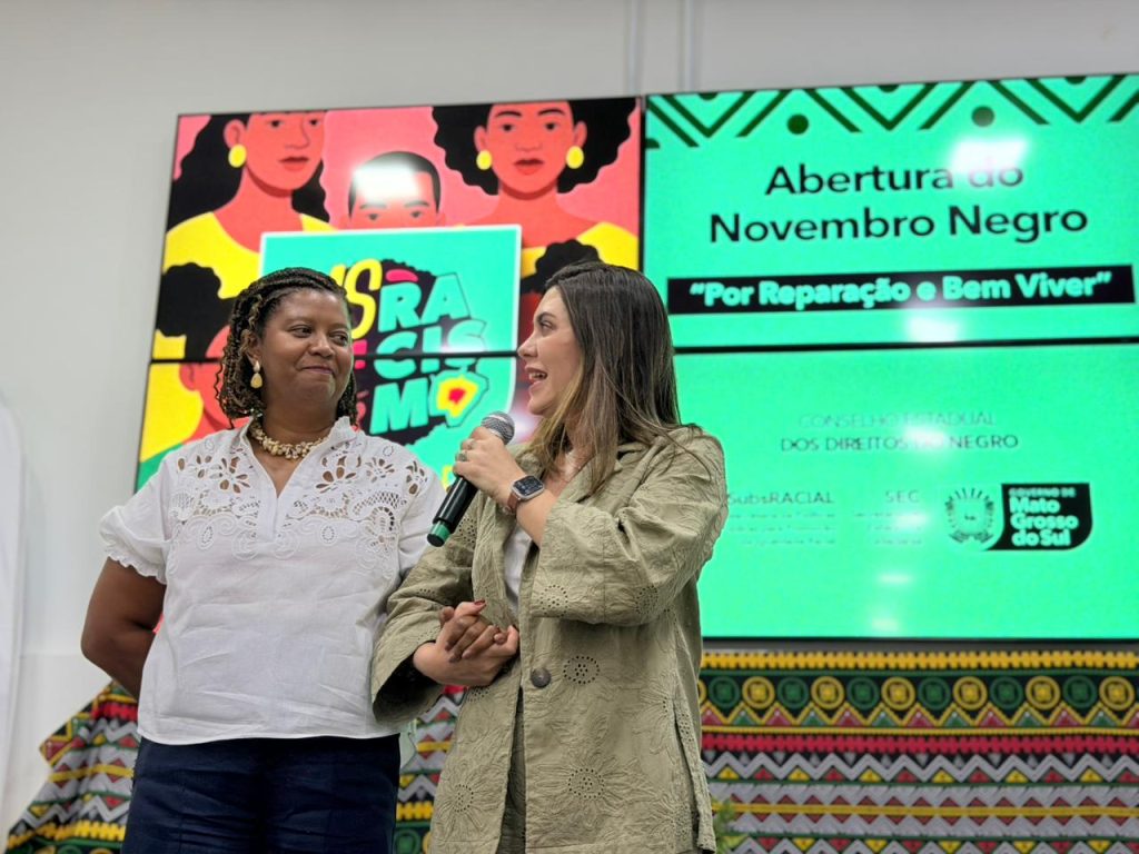 Duas mulheres no palco, uma de blusa branca e outra de casaco caqui segurando um microfone, falam em frente a um telão com o título "Abertura do Novembro Negro".