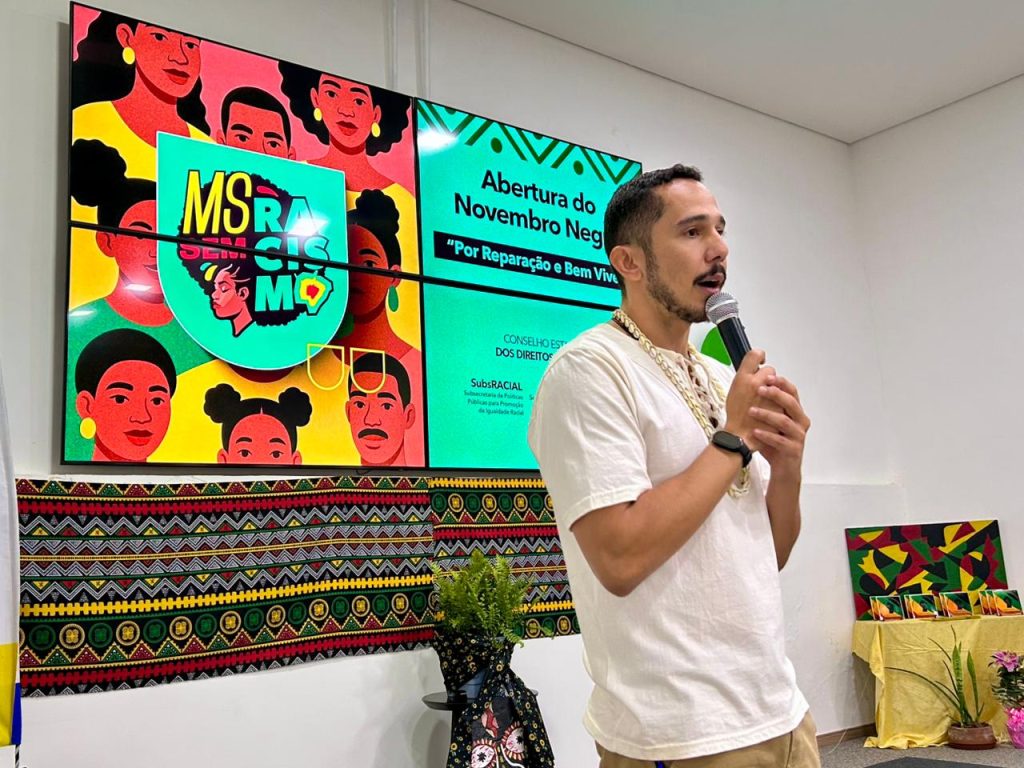 Um homem de camisa branca e colares fala ao microfone, em frente a um telão colorido com o tema "Abertura do Novembro Negro" e a arte da campanha "MS Sem Racismo".