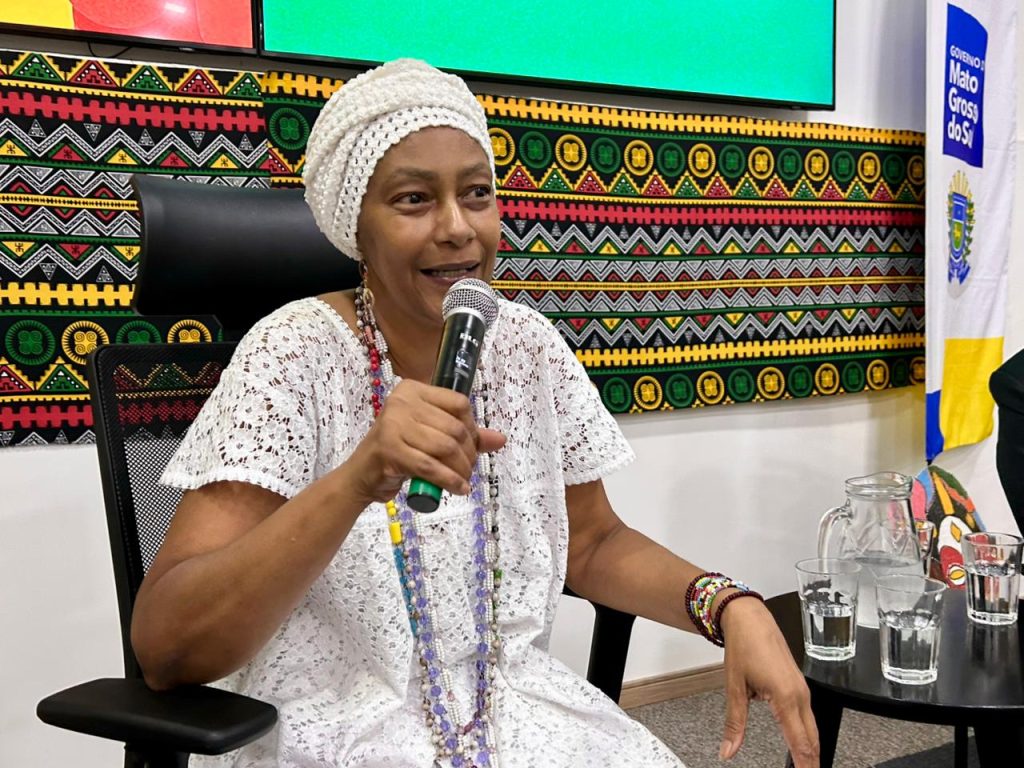 Uma mulher sorrindo, usando turbante branco, vestido de renda e longos colares coloridos, falando ao microfone com um painel de tecido afro-brasileiro ao fundo.