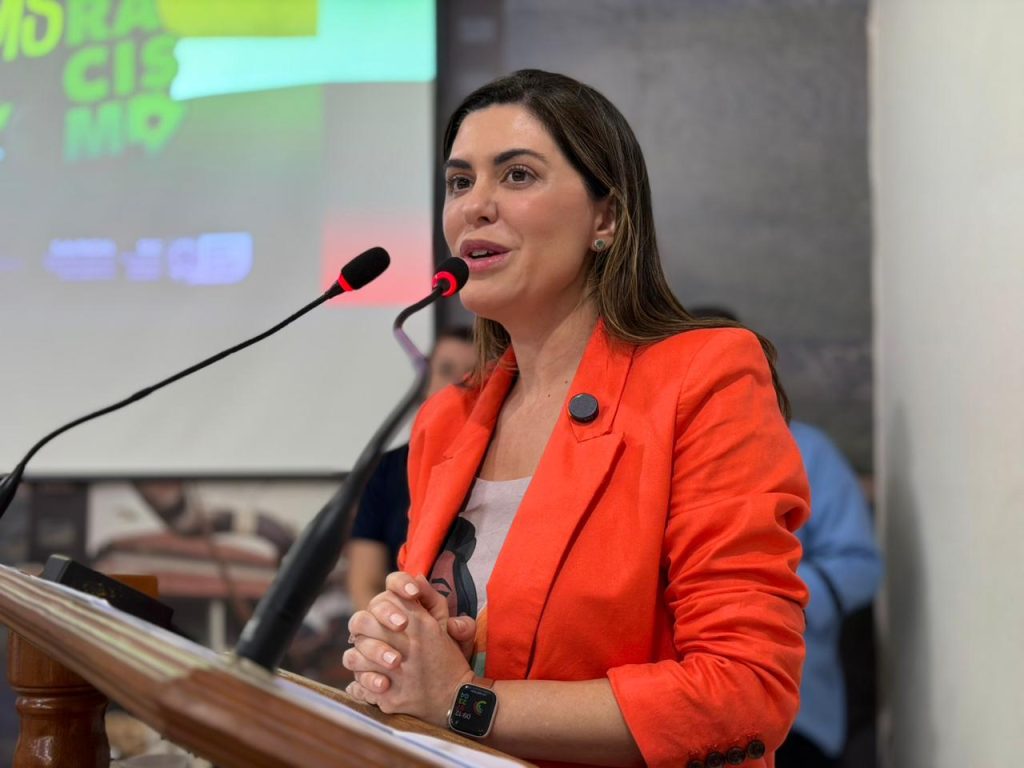 Uma mulher branca com cabelo castanho, vestindo um blazer laranja vibrante no púlpito. Ela está falando em dois microfones, com as mãos unidas e uma tela de projeção com a frase "MS sem Racismo" desfocada ao fundo.