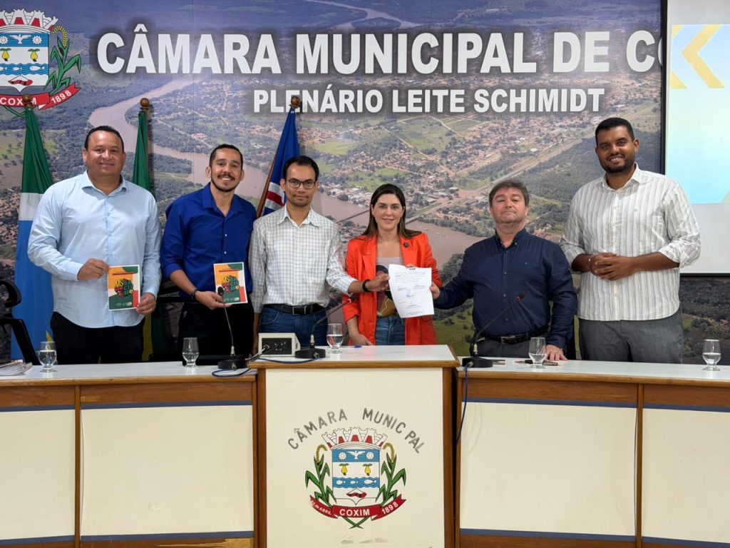 Foto de grupo no plenário, com seis pessoas (quatro homens e uma mulher de blazer laranja, e outro homem) em pé atrás de uma mesa, posando para a foto após a assinatura. A mulher no centro segura um documento assinado, e o fundo é um painel panorâmico da cidade de Coxim.