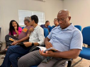 Um homem negro de meia-idade, vestindo uma camisa social azul clara e óculos, está sentado em uma cadeira azul e foca a atenção na leitura do guia MS sem Racismo. Outras pessoas estão sentadas ao fundo, algumas interagindo.