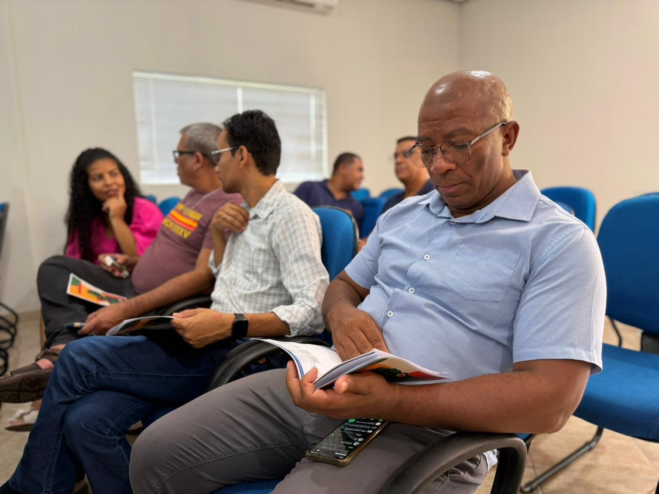 Um homem negro de meia-idade, vestindo uma camisa social azul clara e óculos, está sentado em uma cadeira azul e foca a atenção na leitura do guia MS sem Racismo. Outras pessoas estão sentadas ao fundo, algumas interagindo.