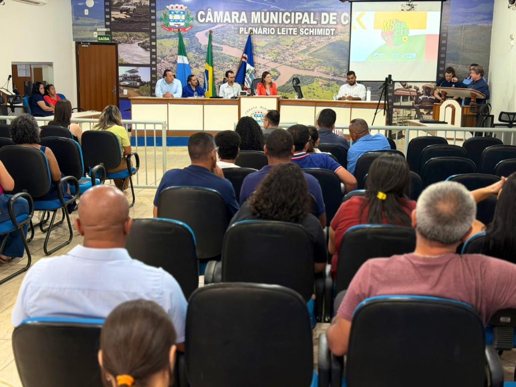 Vista geral do Plenário Leite Schmidt da Câmara Municipal de Coxim, durante o evento. Na mesa diretora, há um grupo de pessoas discursando, enquanto a plateia lotada assiste de frente, vista predominantemente por trás (nuca).