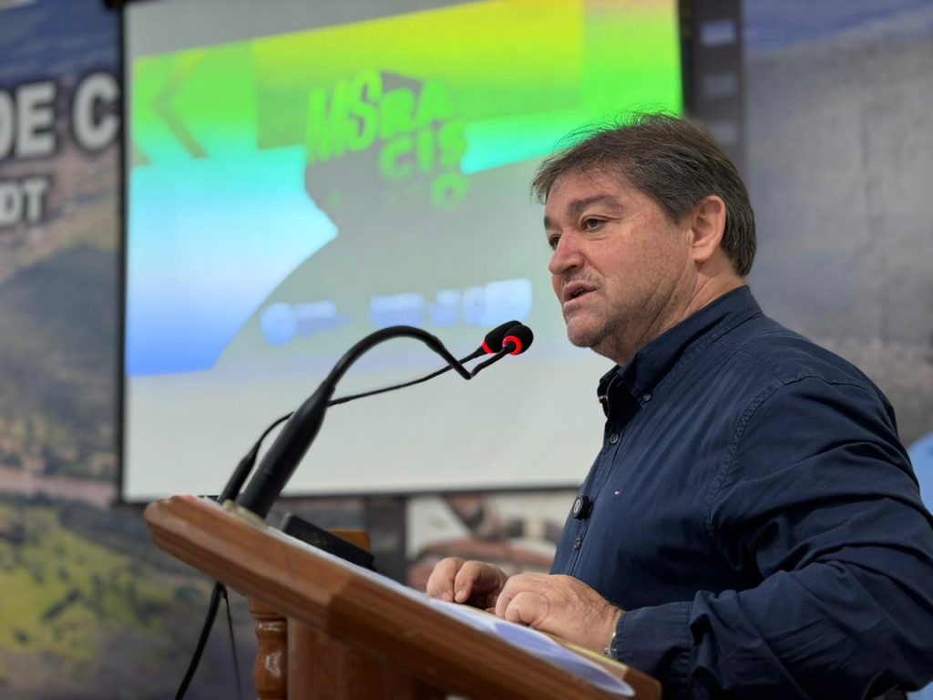 Um homem branco, vestindo uma camisa social azul escura, no pódio. Ele está falando em um microfone com expressão séria, durante o evento.