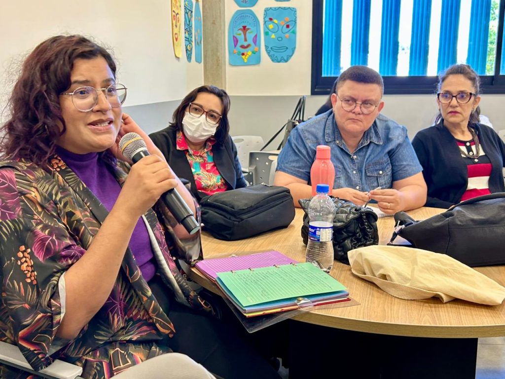 Foto de close de uma mulher morena, à esquerda, usando óculos e um casaco estampado de folhas, falando ativamente em um microfone de mão durante uma mesa de trabalho redonda. Três outros participantes (duas mulheres e um homem de camisa jeans) estão sentados à mesa, acompanhando a fala.