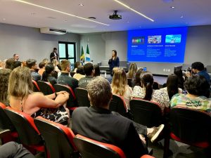 Vista geral de uma audiência em um auditório moderno e escuro, com cadeiras vermelhas. Uma mulher palestrante está em pé na frente (centro), falando para o público. A tela de projeção atrás dela exibe as palavras-chave "GÊNERO", "INTERCULTURALIDADE" e "IDENTIDADE" com pequenos textos explicativos. A plateia é diversificada e inclui pessoas vestindo uniformes de Polícia Militar.