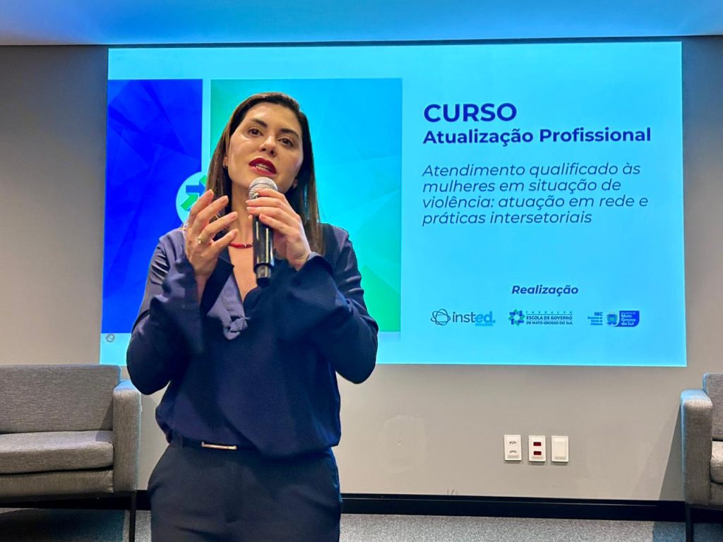 Foto de close de uma mulher em pé, vestindo uma blusa azul-marinho, falando em um microfone de mão com uma expressão séria. Atrás dela, uma tela de projeção exibe o título: "CURSO Atualização Profissional" e o subtítulo: "Atendimento qualificado às mulheres em situação de violência: atuação em rede e práticas intersetoriais". A tela também mostra os logotipos dos realizadores (Insted, Governo do Estado de MS, SEC, Escola de Governo).