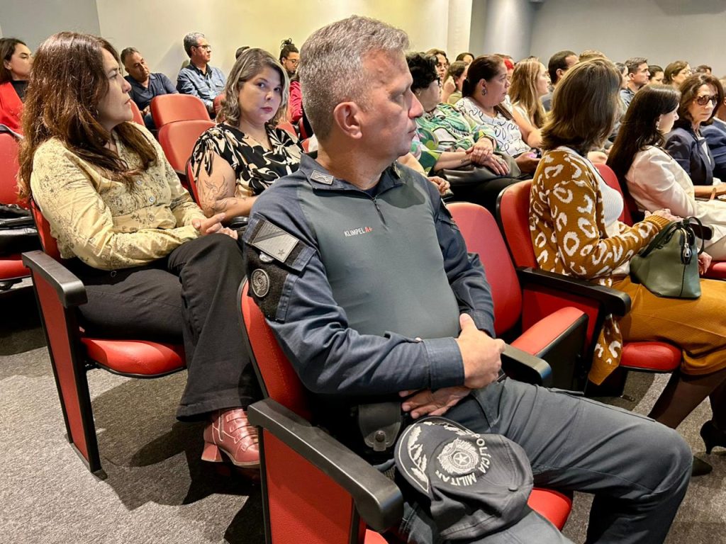 Um homem policial militar (identificado pelo uniforme escuro e a marca "Klimple" e boné na mão), sentado em uma cadeira vermelha em meio à plateia, assistindo atentamente à palestra. Outros participantes estão sentados ao seu redor.