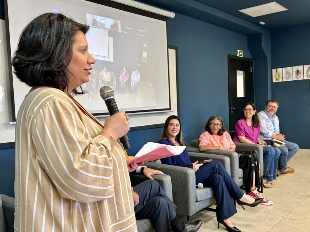 Uma mulher, em primeiro plano à esquerda, vestindo uma blusa listrada bege e branca, está falando com um microfone de mão. Quatro outras pessoas (três mulheres e um homem) estão sentadas em cadeiras cinzas em segundo plano, formando uma mesa de debate. Atrás, uma tela de projeção branca exibe uma transmissão online.