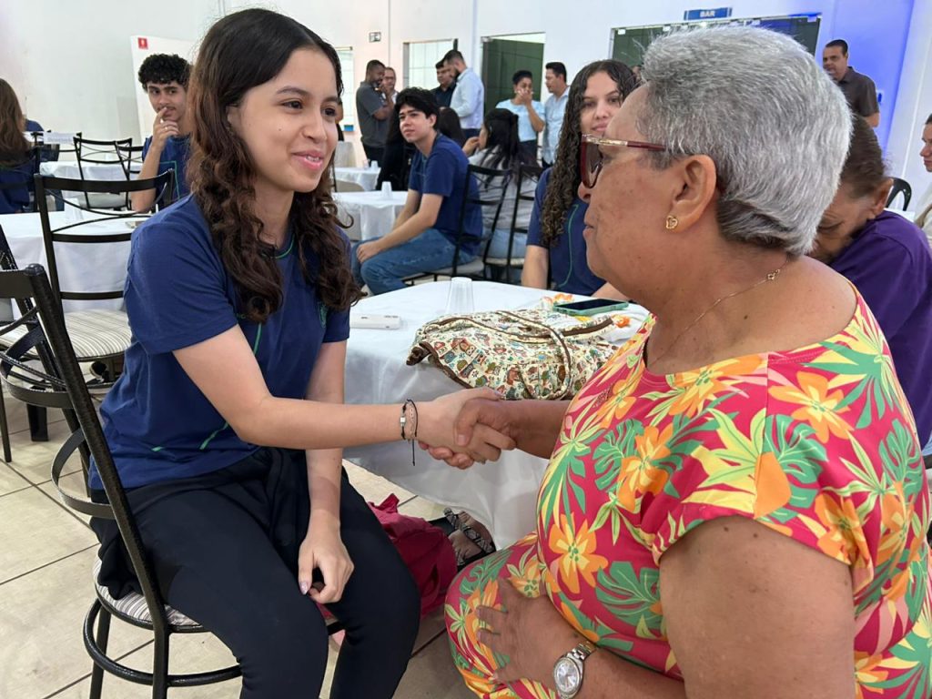 Em primeiro plano, uma jovem de cabelo cacheado e camiseta azul (à esquerda) aperta a mão de uma mulher idosa (à direita). A mulher idosa, de sorriso aberto, usa um vestido florido nas cores laranja, amarelo e verde. Ambas estão sentadas à mesa, num gesto de cordialidade e conexão.