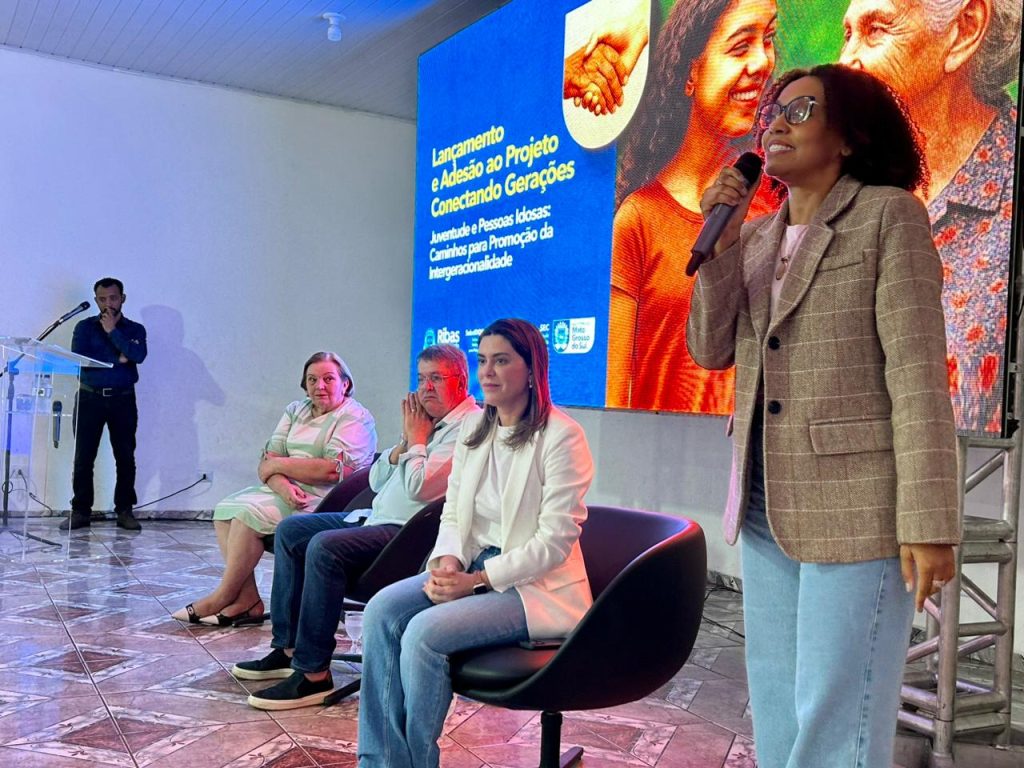 Em primeiro plano e de pé no lado direito, uma mulher de pele negra, blazer xadrez e calça jeans fala ao microfone. Atrás dela, no painel digital iluminado, lê-se: "Lançamento e Adesão ao Projeto Conectando Gerações: Juventude e Pessoas Idosas: Caminhos para Promoção da Intergeracionalidade." Sentados em cadeiras no palco, à esquerda, estão três pessoas: uma mulher idosa de vestido claro, um homem de camisa azul clara e barba, e uma mulher jovem de blazer branco.