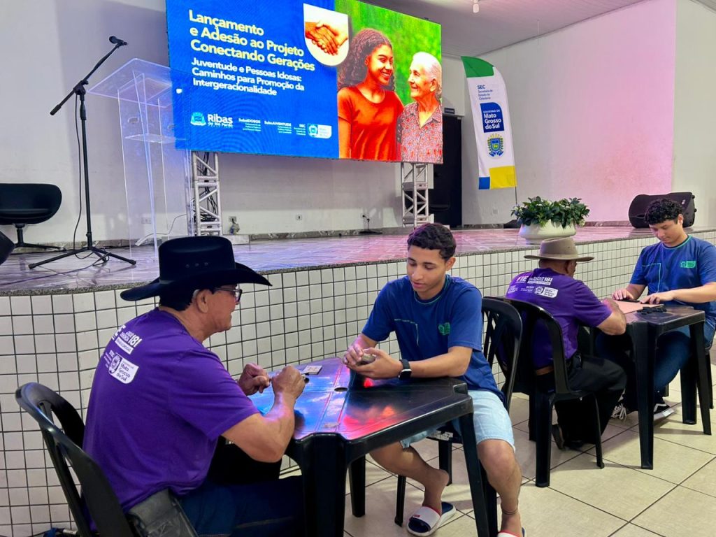 Pessoas jogando baralho em uma mesa escura. No primeiro plano, um homem idoso de chapéu de cowboy preto e camiseta roxa está sentado, jogando cartas com um jovem de camiseta azul, ambos concentrados no jogo. Ao fundo, em outra mesa, outros dois jovens, também de camiseta azul, jogam. A cena mostra a integração através de atividades recreativas. O telão ao fundo confirma o nome do projeto.