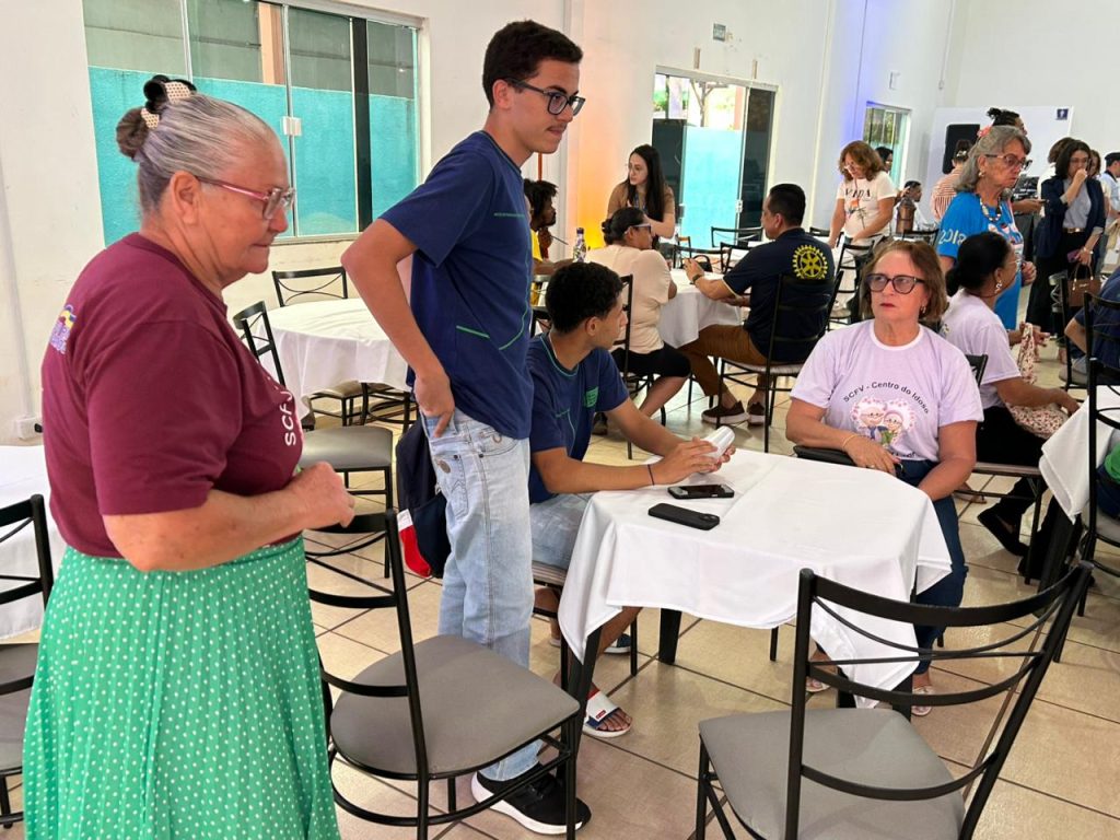 No primeiro plano, à esquerda, uma mulher idosa de cabelo preso, óculos, camiseta vinho e saia verde de bolinhas, está de pé conversando com um jovem alto de camiseta azul escura. Outras duas pessoas estão sentadas à mesa: uma mulher de óculos e camiseta branca, e um jovem manuseando um objeto na mão (possivelmente outro celular ou cartão). A cena capta a troca entre as gerações.