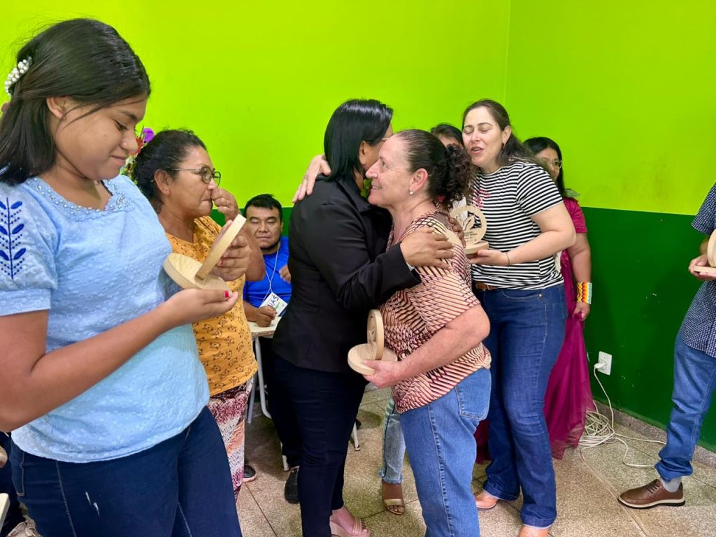 Momento de abraço e celebração. Uma mulher de preto abraça outra mulher com uma blusa listrada bege e preta. Mulheres indígenas ao redor seguram e admiram troféus do evento.
