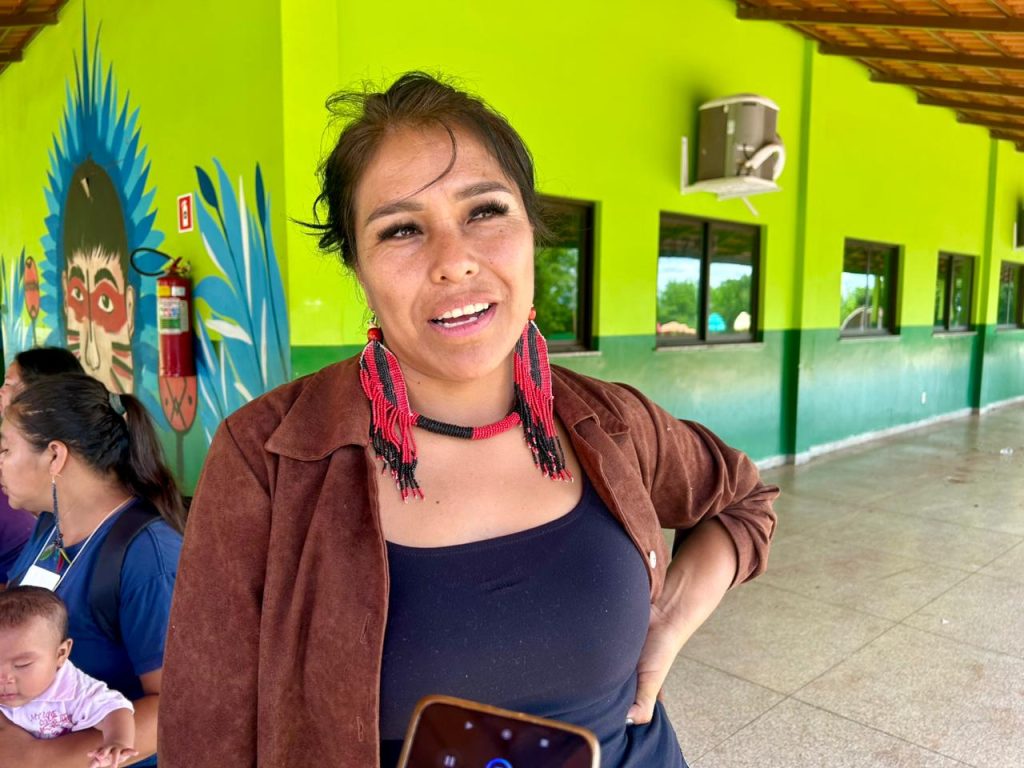 Uma mulher indígena sorrindo e olhando para cima, com brincos e colar de contas pretas e vermelhas longos. Ela veste uma blusa preta e um casaco marrom, e está em frente a uma parede verde com uma pintura mural.