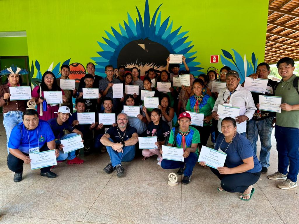 Foto de grupo de aproximadamente 30 participantes, a maioria indígena, em frente a uma parede verde com grafismos artísticos em azul e preto. Todos seguram certificados do curso Empretec. Há homens e mulheres, alguns trajados com adornos tradicionais, celebrando o encerramento do programa.