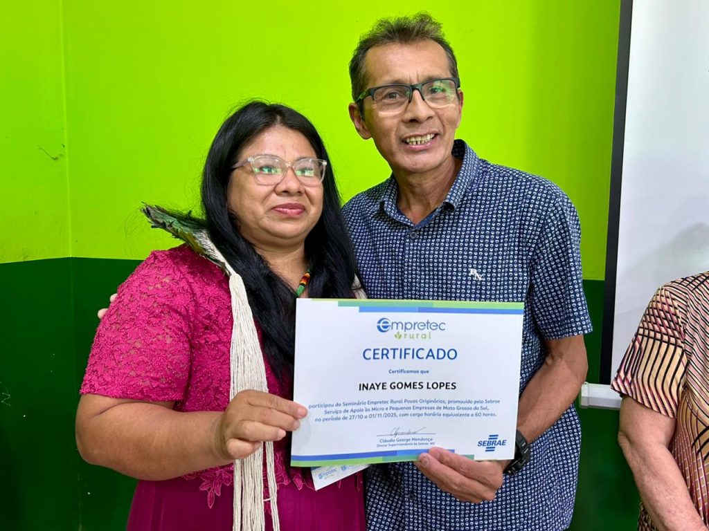 Uma mulher indígena usando um vestido rosa e um adorno com penas, sorri enquanto segura seu certificado do "Empretec Rural Povos Originários". Ao seu lado, um homem sorri. O certificado é do SEBRAE e indica a participação de 27/10 a 01/11/2023.
