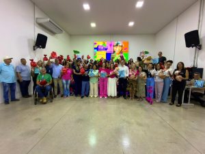 Uma foto de grupo tirada de longe que mostra dezenas de pessoas, incluindo autoridades e voluntários, posando em uma sala de evento. O grupo está organizado em várias filas, todos sorrindo e segurando pacotes de presente, simbolizando o sucesso da campanha "Caixa Encantada".