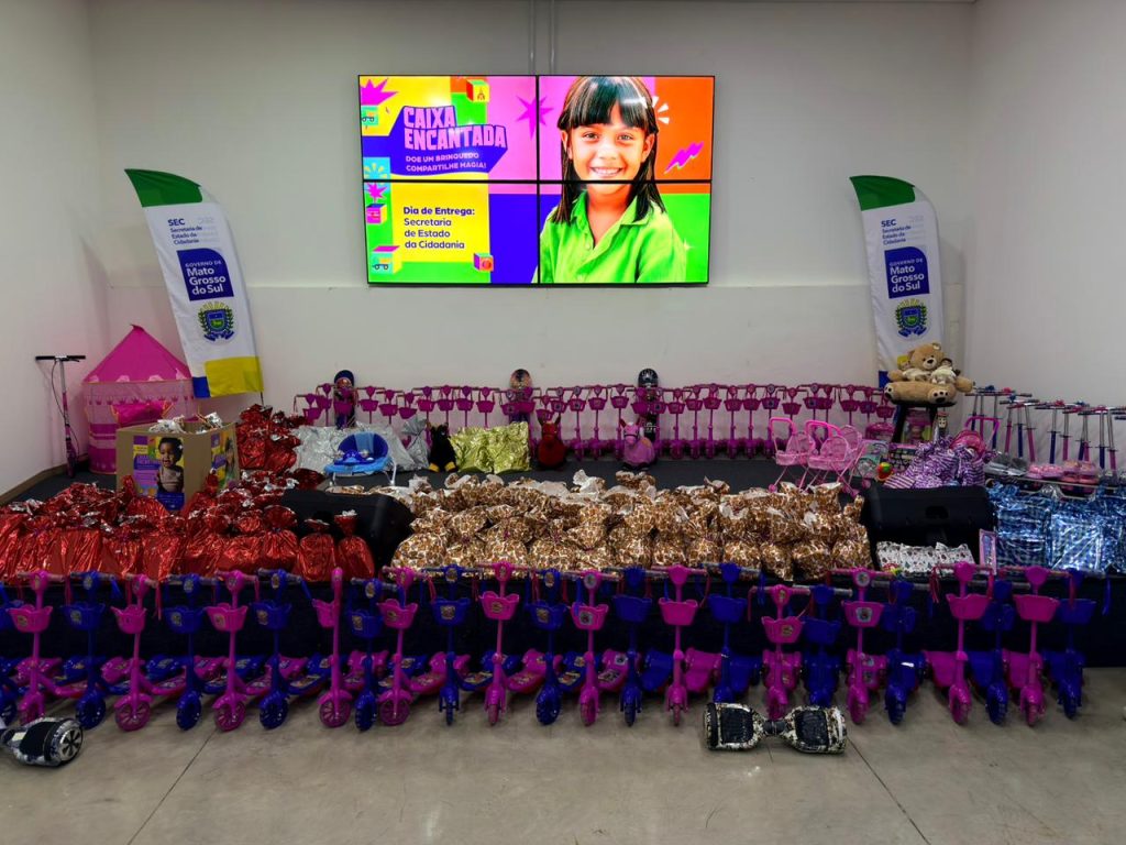 Uma visão geral de um grande salão com uma enorme quantidade de brinquedos e presentes prontos para doação. No centro, há várias fileiras de patinetes de três rodas em cores vibrantes, rosa choque e azul, totalizando dezenas de unidades. Na frente e ao lado, pilhas de pacotes embrulhados em papel de presente vermelho e estampado (laranja e branco). No fundo, um grande painel de LED exibe o logo da campanha "CAIXA ENCANTADA" com o lema "DOE UM BRINQUEDO COMPARTILHE MAGIA!" e a imagem de uma menina sorrindo. Estão presentes também a bandeira e o logo da SEC do Governo do Estado de Mato Grosso do Sul.