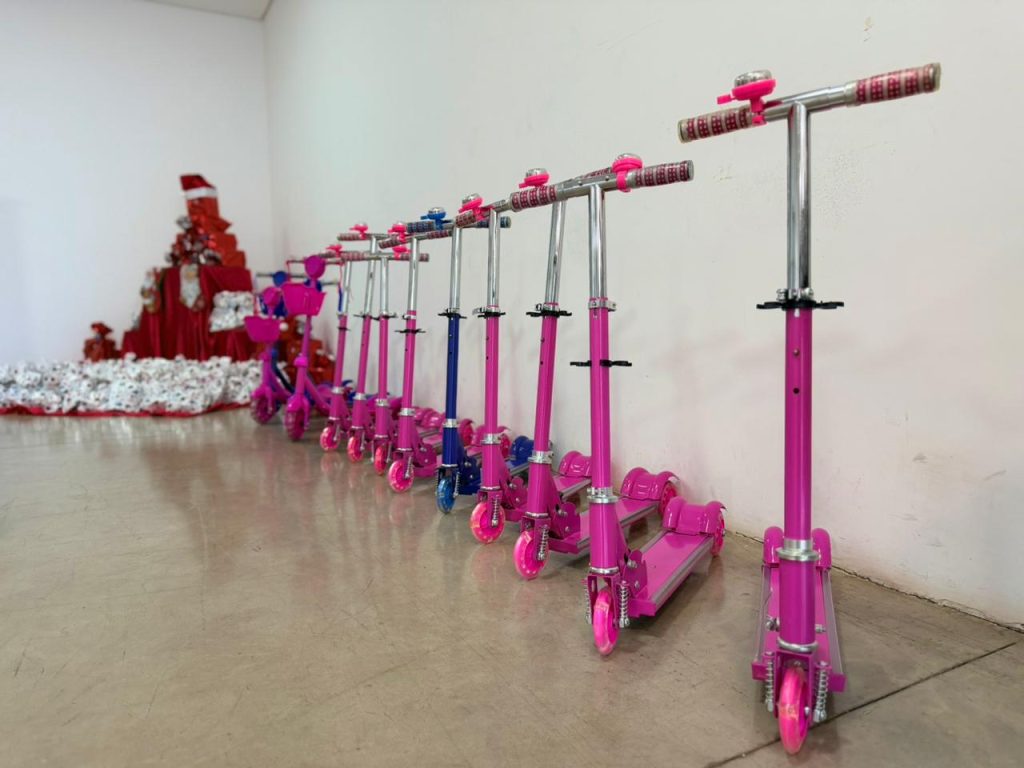 Uma fileira de 10 patinetes de três rodas, predominantemente na cor rosa choque, alinhados no centro de um piso de cimento. Cada patinete tem detalhes cromados e pequenos sinos rosa nas hastes do guidão. No fundo esquerdo, fora de foco e contra uma parede branca, há uma grande montanha de presentes embrulhados em papel estampado e uma estrutura que lembra uma árvore de Natal feita de caixas vermelhas. O cenário mostra a doação de brinquedos.