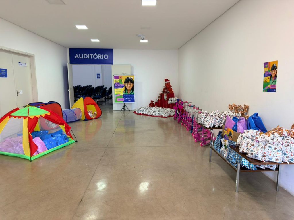 Uma foto de ambiente que mostra a organização do local de entrega dos presentes. À esquerda, encostada na parede, há uma barraca infantil colorida em formato de túnel. Mais ao fundo, na parede branca, uma placa indica "AUDITÓRIO" com setas para dentro. No lado direito do salão, estão dispostos montes de presentes embrulhados, e em segundo plano, uma fileira de patinetes rosa. Um cartaz da campanha "Caixa Encantada" é visível na parede central.