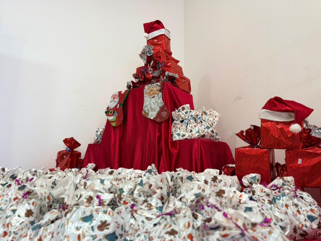 Uma pilha central de presentes com tema natalino. No primeiro plano, uma grande quantidade de pacotes embrulhados em papel de presente branco com estampas coloridas (corações, árvores, doces) está espalhada pelo chão. Atrás, uma mesa coberta com tecido de veludo vermelho sustenta uma pilha de caixas maiores, todas embrulhadas em papel vermelho metálico e empilhadas de forma a criar uma "árvore de Natal" simbólica. Meias de Natal decorativas pendem do tecido vermelho.
