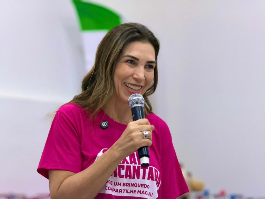 Primeira-Dama de Mato Grosso do Sul, Mônica Riedel, está sorrindo, virada para a direita, e segurando um microfone perto da boca, indicando que está falando com o público. Ela usa a camiseta rosa vibrante da campanha, que mostra parcialmente o texto "CAIXA ENCANTADA - DOE UM BRINQUEDO COMPARTILHE MAGIA!". Ela tem cabelos castanhos longos e usa um anel grande na mão que segura o microfone. 