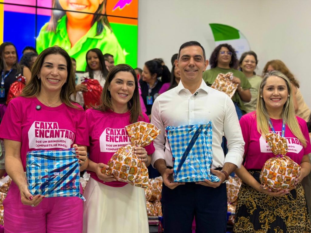 Quatro pessoas adultas, duas mulheres e dois homens, sorrindo e posando para a foto em primeiro plano, segurando presentes embrulhados. As duas mulheres nas pontas usam camisetas rosa com a estampa "CAIXA ENCANTADA - DOE UM BRINQUEDO COMPARTILHE MAGIA!". A mulher à esquerda segura um pacote azul xadrez, e a mulher à direita um pacote estampado em tons de laranja. O homem no centro, de camisa social branca, e a mulher ao lado dele, de camiseta rosa, também seguram pacotes embrulhados. Atrás deles, um grupo de pessoas igualmente sorridentes, muitas vestindo camisetas do evento, e um telão digital no fundo com imagens coloridas.
