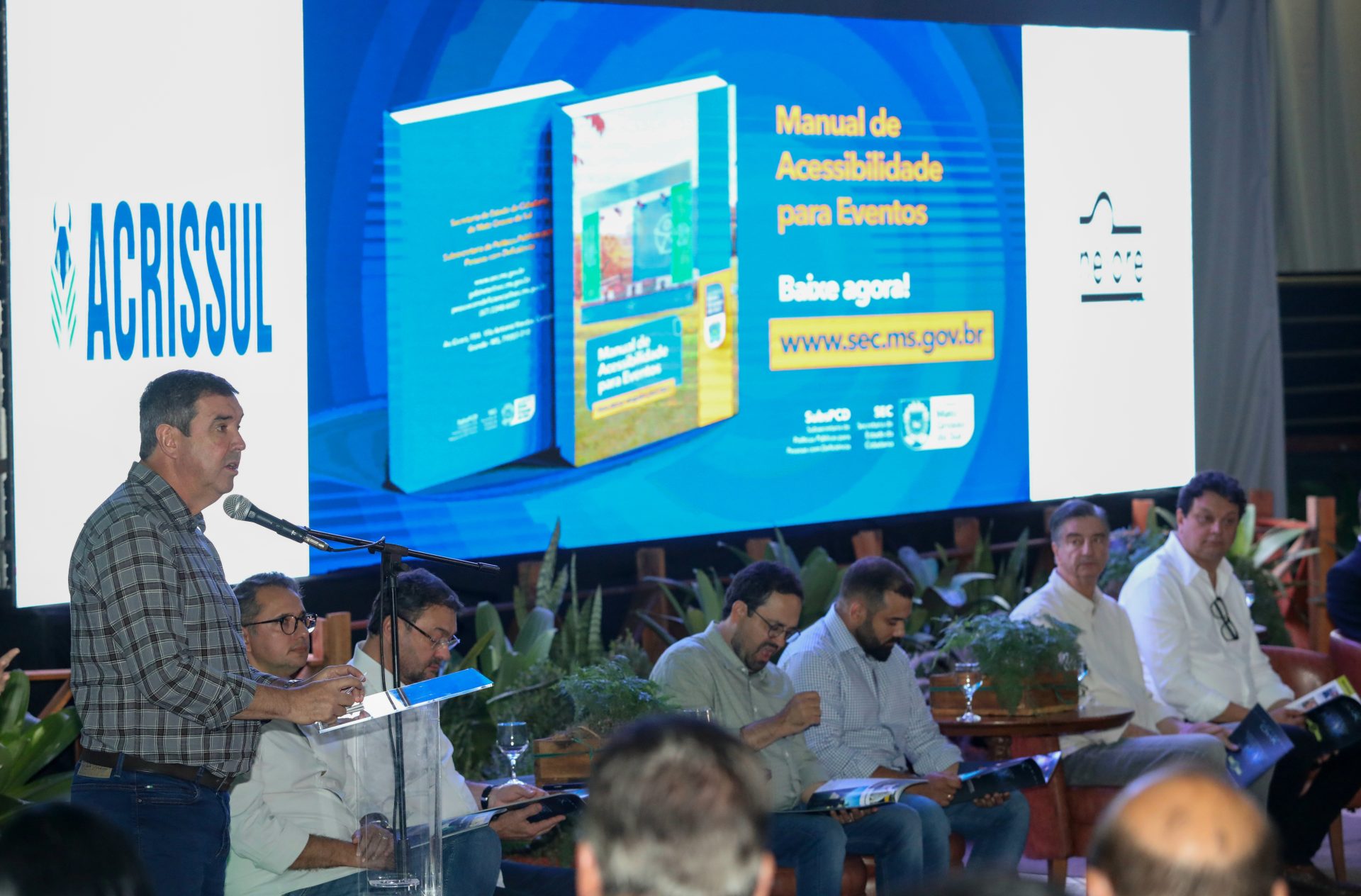 Governador do Estado, Eduardo Riedel, veste camisa xadrez e calça jeans, está de pé à esquerda, falando ao microfone em um púlpito com uma pasta de documentos azul. Ele se dirige a uma plateia de homens sentados em cadeiras estofadas, alguns segurando cadernos ou tablets. O fundo da cena é dominado por um grande painel de LED azul. O painel exibe a capa de um livro intitulado "Manual de Acessibilidade para Eventos" e uma instrução para baixá-lo no site oficial, além do logo da ACRISSUL e outros logos institucionais. O cenário é formal, sugerindo o lançamento do evento.