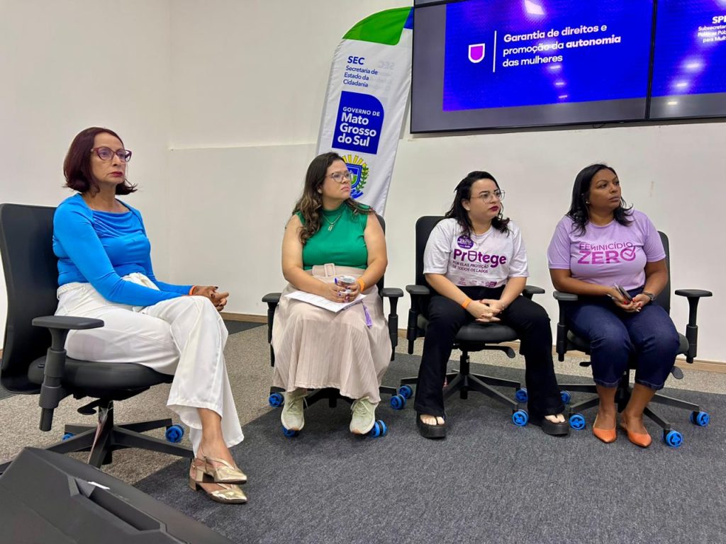 Quatro mulheres sentadas em cadeiras giratórias pretas, formando uma mesa de debate. Elas estão voltadas para o lado esquerdo da imagem, atentas. Um cartaz ao fundo exibe a frase "Garantia de direitos e promoção da autonomia das mulheres" e a bandeira do Governo do Estado de Mato Grosso do Sul.