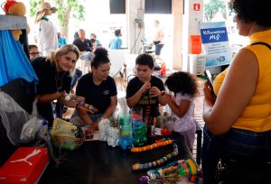 Uma cena em uma tenda de eventos mostrando uma oficina de artesanato e sustentabilidade. Quatro pessoas estão inclinadas em torno de uma mesa preta cheia de materiais de reciclagem, como garrafas plásticas coloridas, tampas, tecidos e tubos de papelão decorados. Uma mulher de cabelos loiros e camiseta escura sorri para a câmera enquanto segura um artesanato colorido. Ao lado dela, outra mulher e um adolescente, ambos de camisetas escuras, trabalham em projetos. Uma menina de cabelos cacheados está concentrada em seu trabalho. Os itens na mesa sugerem a confecção de brinquedos a partir de lixo reciclado. No fundo, um banner vertical azul exibe o logo da campanha "Sustentabilidade MS" do Governo do Estado de Mato Grosso do Sul.