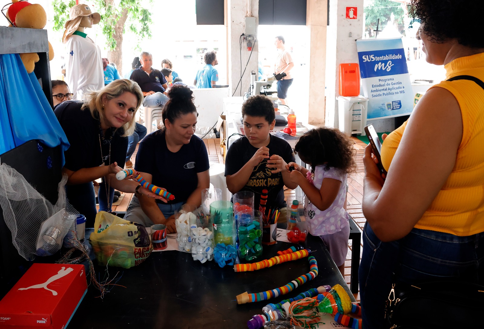 Uma cena em uma tenda de eventos mostrando uma oficina de artesanato e sustentabilidade. Quatro pessoas estão inclinadas em torno de uma mesa preta cheia de materiais de reciclagem, como garrafas plásticas coloridas, tampas, tecidos e tubos de papelão decorados. Uma mulher de cabelos loiros e camiseta escura sorri para a câmera enquanto segura um artesanato colorido. Ao lado dela, outra mulher e um adolescente, ambos de camisetas escuras, trabalham em projetos. Uma menina de cabelos cacheados está concentrada em seu trabalho. Os itens na mesa sugerem a confecção de brinquedos a partir de lixo reciclado. No fundo, um banner vertical azul exibe o logo da campanha "Sustentabilidade MS" do Governo do Estado de Mato Grosso do Sul.