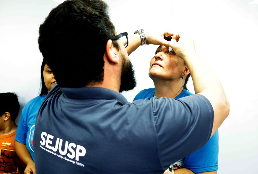 Em destaque, um profissional de saúde realizando um exame oftalmológico rápido em uma mulher. O profissional, um homem barbudo de camisa azul-marinho com o logo "SEJUSP" nas costas, está pingando colírio ou examinando o olho de uma mulher. A mulher está com o rosto inclinado para cima e os olhos abertos. A luz de fundo é forte e branca, destacando a ação. A imagem ilustra a prestação de serviços de saúde.