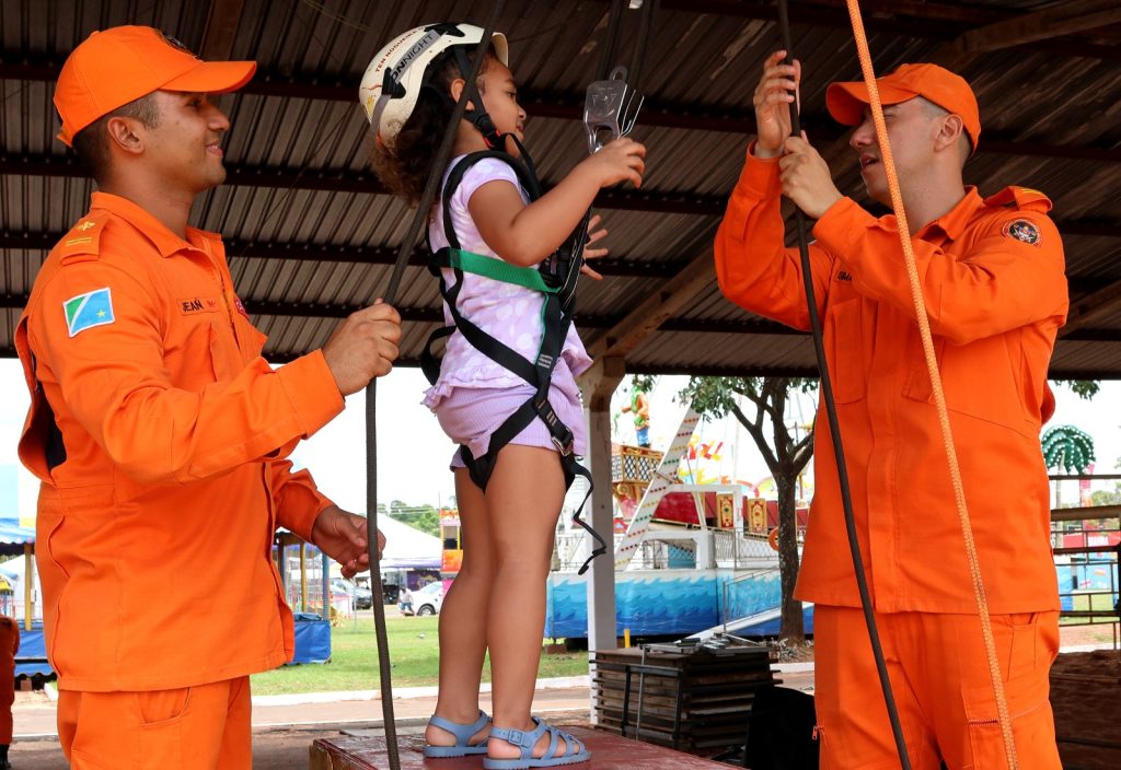 Uma foto de dois bombeiros em uniformes laranjas e bonés, auxiliando uma criança pequena a se preparar para uma atividade de aventura: tirolesa. A menina está de pé em uma plataforma de madeira, usando um capacete de segurança branco e um cinto de segurança (baudrier) preto. Os dois bombeiros seguram cordas e estão checando o equipamento de segurança da criança, que segura as alças do cinto com as mãos. A cena é coberta por um telhado de metal e, ao fundo, é possível ver as estruturas de um parque de diversões.