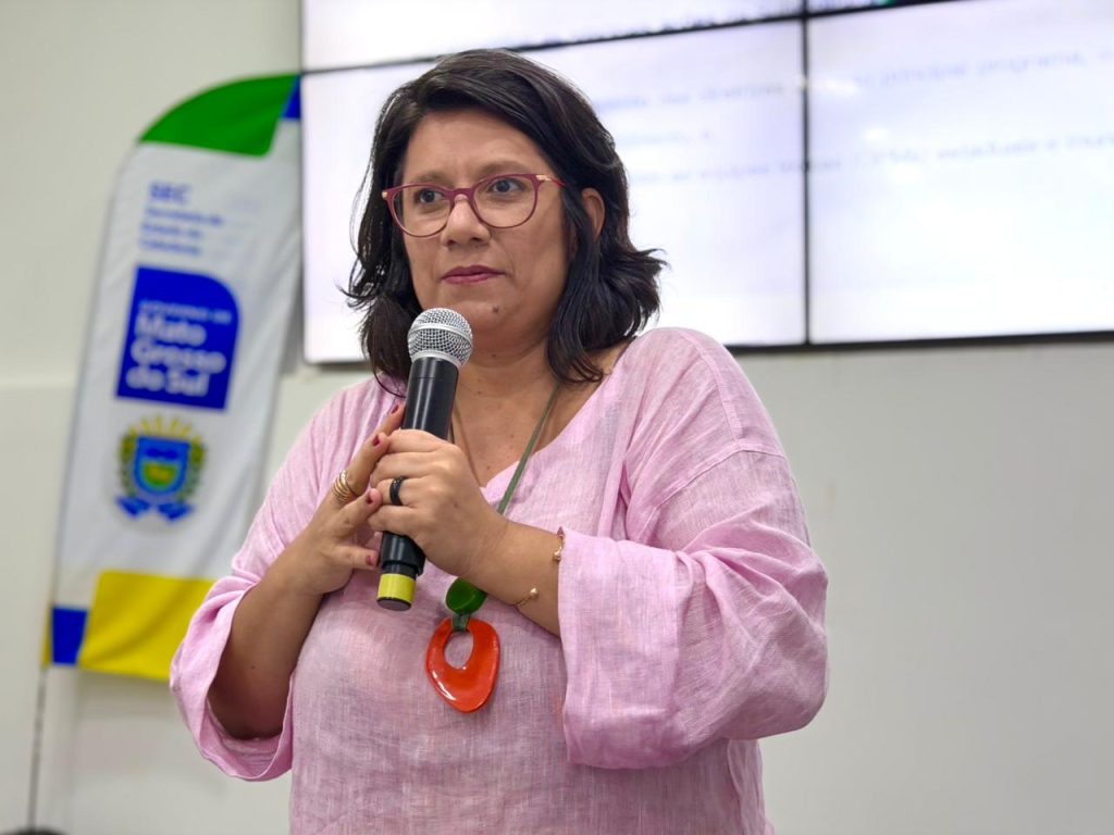 Foto de close de uma mulher de óculos e cabelo escuro, vestindo uma blusa rosa claro e um colar com pingente laranja. Ela está em pé, segurando um microfone com ambas as mãos e falando. Ao fundo, parcialmente visível, há uma bandeira da Secretaria de Estado de Cidadania (SEC) de Mato Grosso do Sul.
