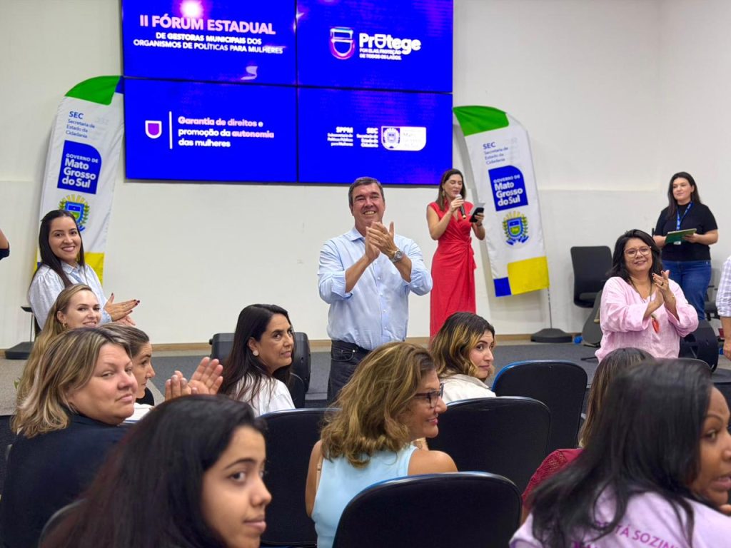Foto de um momento de aplausos. Uma plateia majoritariamente feminina é vista em primeiro plano, aplaudindo. No centro, um homem em camisa azul clara e uma mulher em vestido vermelho, em segundo plano, também aplaudem. O painel de LED ao fundo exibe o título do fórum e a frase "Garantia de direitos e promoção da autonomia das mulheres".