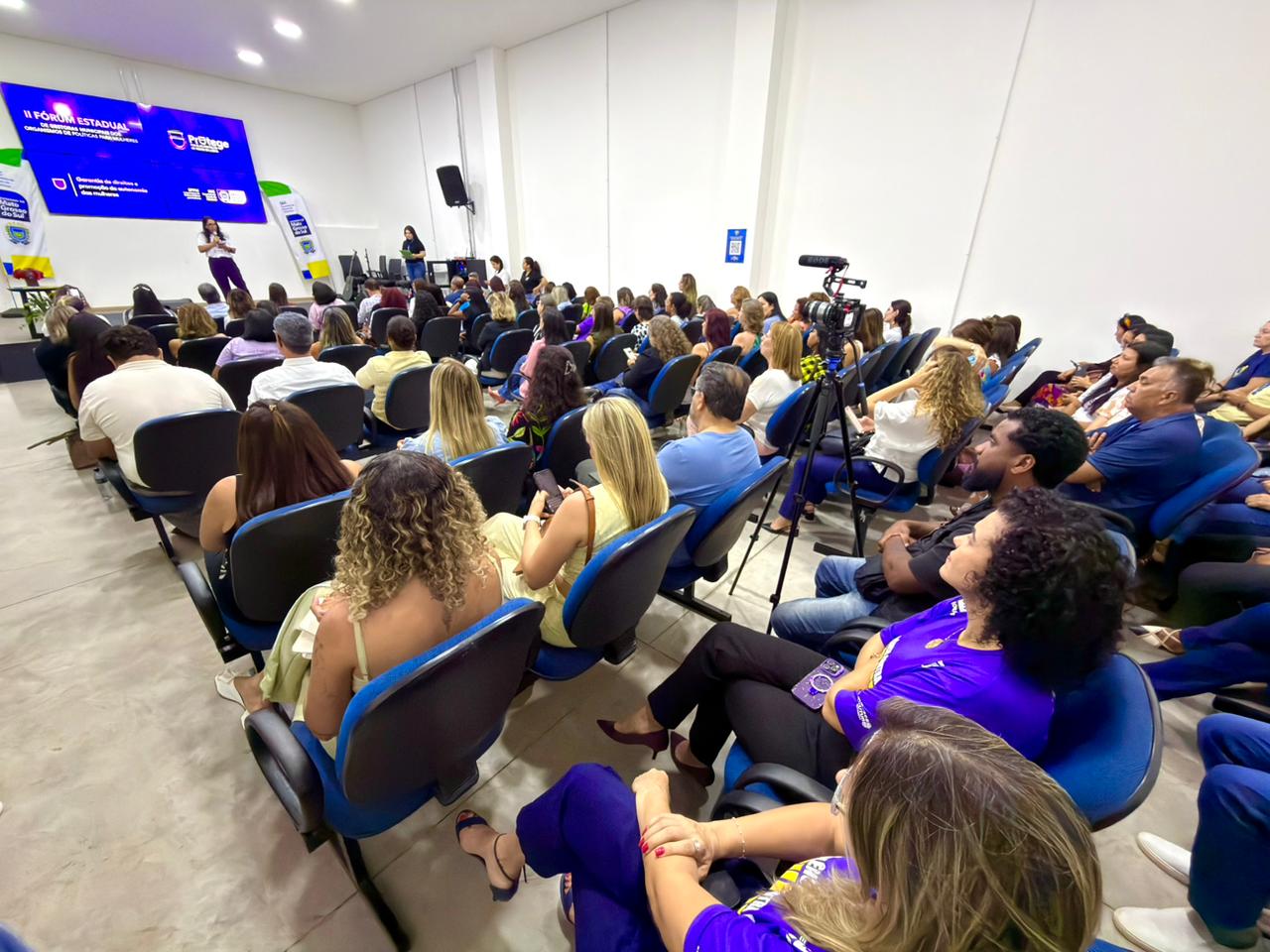 Vista geral do auditório, com uma grande plateia sentada em cadeiras azuis, vista predominantemente por trás. Duas mulheres estão de pé na frente, falando para o público. Um tripé de câmera de vídeo está visível à direita. O telão de projeção ao fundo mostra o título "II Fórum Estadual de Gestoras..." e logotipos.