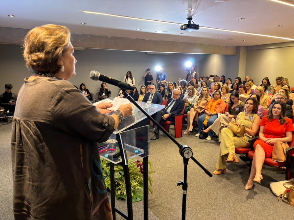 Uma mulher mais velha, de costas para a câmera, com cabelo curto castanho-claro e vestindo uma blusa/túnica marrom e escura com estampa colorida na barra, fala em um púlpito transparente, segurando um papel e um microfone. Ela está em uma posição elevada, dirigindo-se à plateia sentada à frente e ao lado, capturando um momento de discurso e interação.