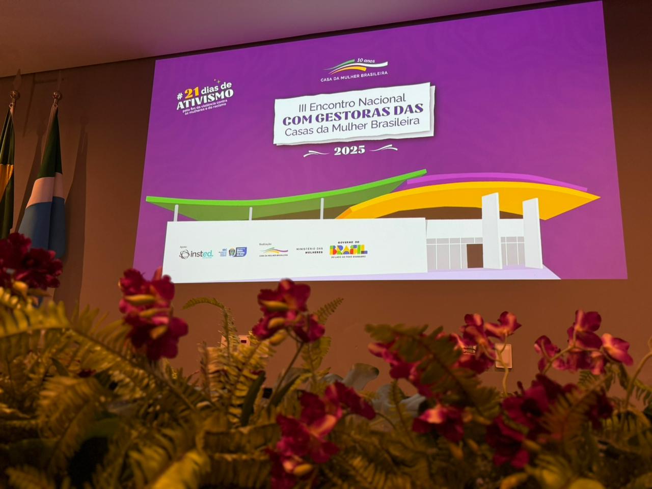 Uma foto em primeiro plano de um grande arranjo floral vermelho e verde, com foco no fundo. O telão projeta a identidade visual do evento em cores vibrantes, principalmente roxo e verde-limão. No centro, o texto: "III Encontro Nacional com Gestoras das Casas da Mulher Brasileira 2025". Abaixo, vê-se um desenho estilizado de uma estrutura que representa a Casa da Mulher Brasileira e logotipos de parceiros e do Governo Federal.