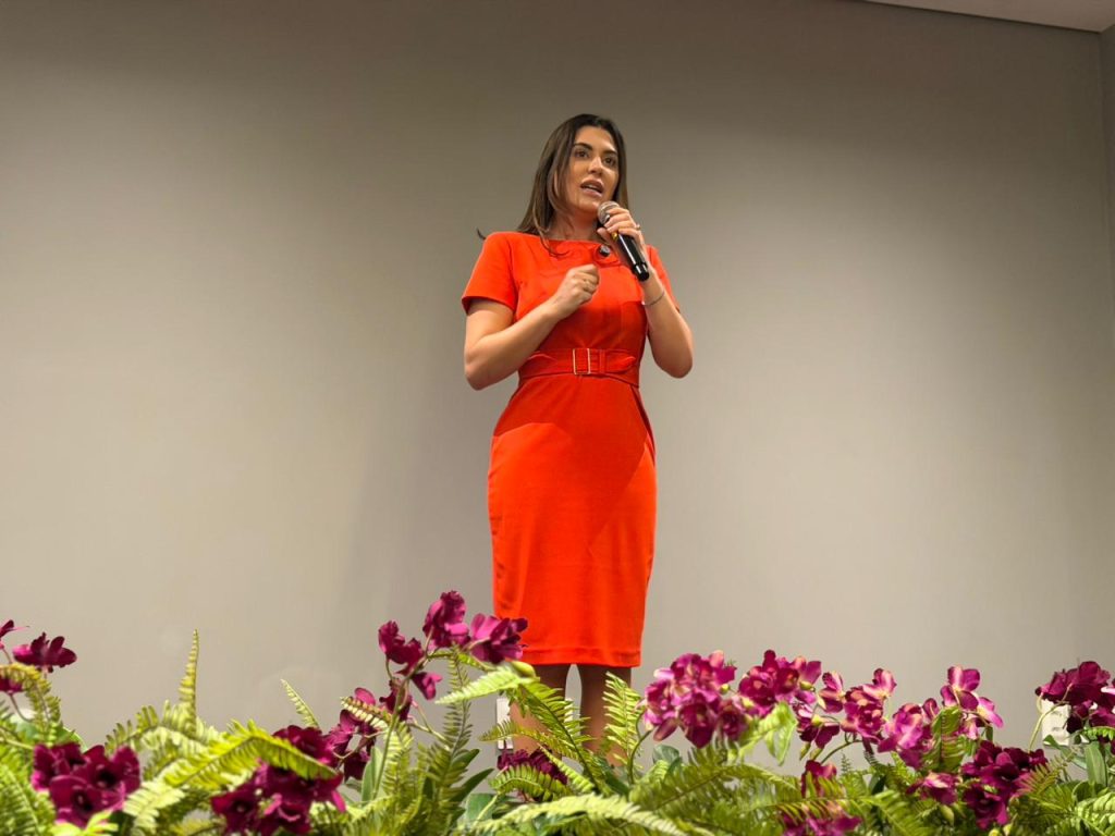 Uma mulher com cabelo castanho escuro e um vestido tubinho laranja de manga curta e cinto fala em pé no palco, segurando um microfone. Ela está em frente a uma parede cinza clara, lisa e sem adornos, e o primeiro plano é dominado pelo mesmo arranjo floral verde e roxo. Sua expressão é focada e engajada com o público.