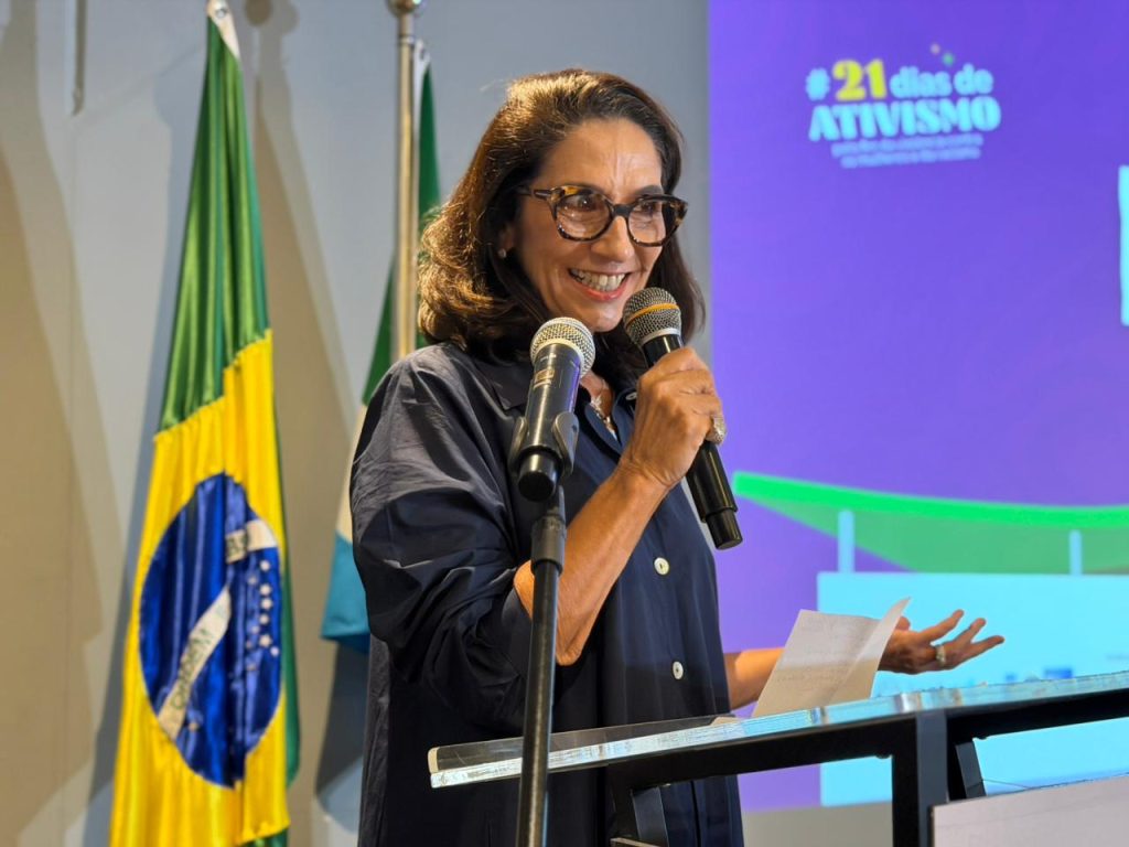 Uma mulher com cabelo castanho e óculos fala ao microfone, posicionada atrás de um púlpito de acrílico. Ela veste uma camisa/bata azul-marinho de botões. Atrás dela, visíveis, estão as bandeiras do Brasil e de um estado (parcialmente visível), e o telão com o texto "#21 dias de ATIVISMO". Ela segura um papel na mão esquerda e sorri levemente enquanto fala.
