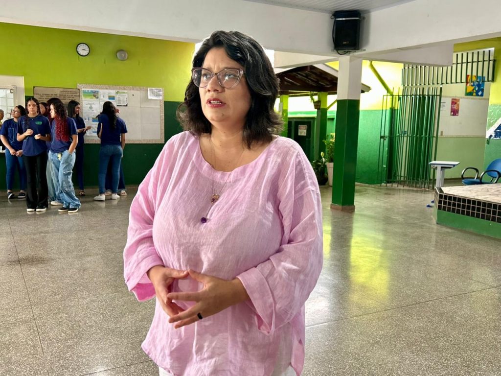 Uma mulher de óculos, cabelos castanhos curtos e blusa rosa clara está em pé no centro de um pátio escolar. Ela esta concedendo uma entrevista. Ao fundo, de forma levemente desfocada, estudantes conversam e a arquitetura da escola mostra colunas verdes e áreas de convivência.