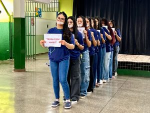 Fotografia em plano médio de uma fila de aproximadamente dez estudantes em um pátio escolar. Elas estão posicionadas de perfil, uma atrás da outra. Todas usam camisetas azuis escuras e possuem um pedaço de fita adesiva branca cobrindo a boca, simbolizando o silenciamento. A primeira jovem da fila segura um cartaz branco com os dizeres em letras vermelhas: “TROCA ESSE SHORT”. Ao fundo, vê-se a estrutura da escola com colunas verdes e grades.