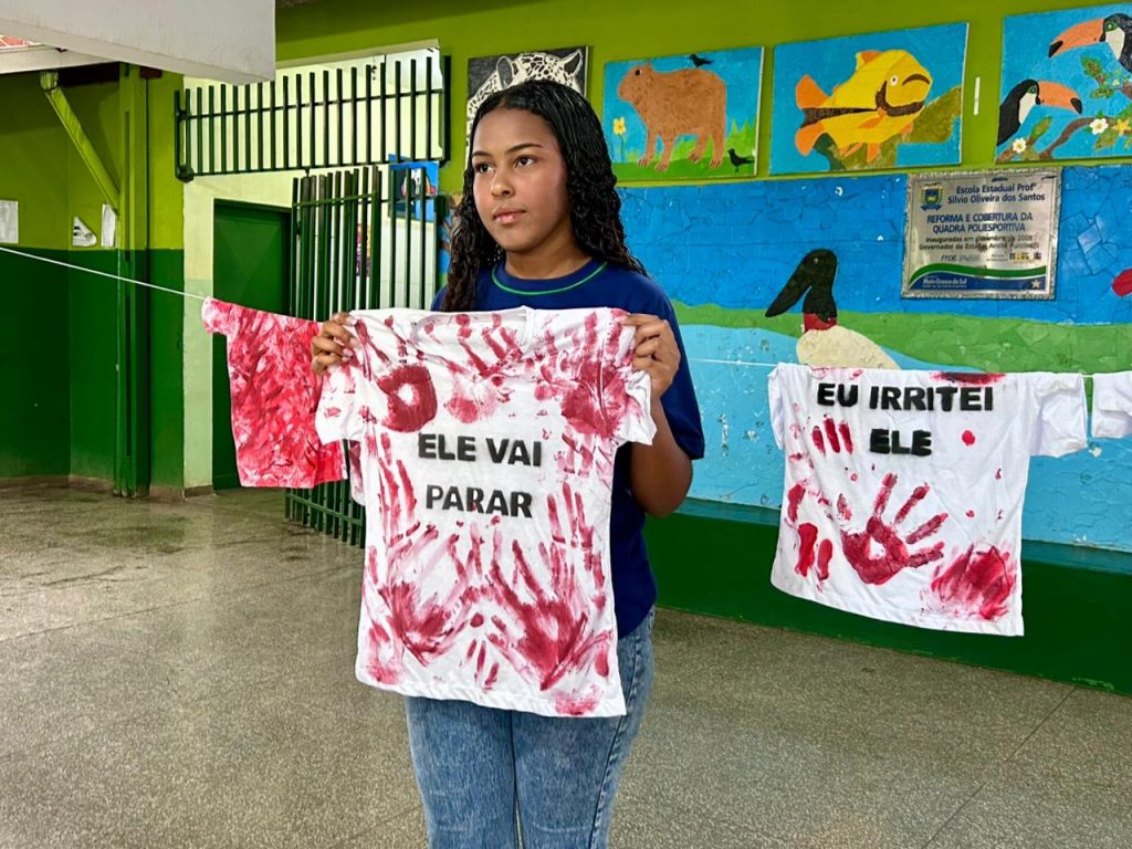 Uma jovem estudante de cabelos cacheados segura à frente do corpo uma camiseta branca estendida. A camiseta está manchada com várias marcas de mãos em tinta vermelha, simulando sangue. No centro da peça, lê-se a frase em letras pretas: “ELE VAI PARAR”. Ao fundo, há um varal com outras camisetas semelhantes e uma parede colorida com pinturas de animais da fauna pantaneira, como uma capivara e um tuiuiú.