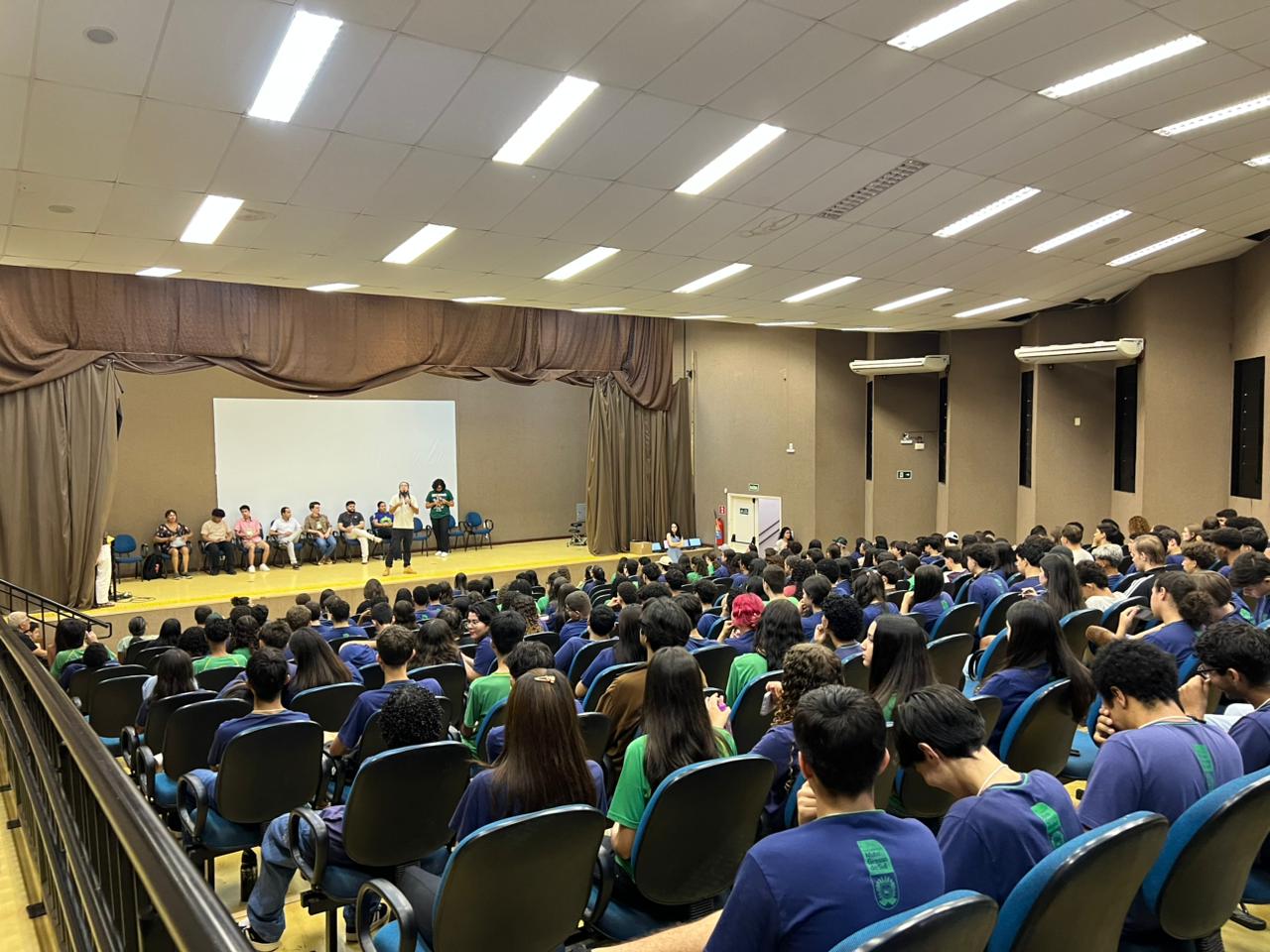A imagem mostra um grande auditório lotado de jovens que veste camisetas nas cores azul-marinho e verde, sentado em cadeiras azuis escuras. Um painel de palestrantes senta-se no palco em uma bancada.